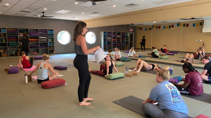 Students lie or sit on their yoga mats in a studio while their professor speaks.
