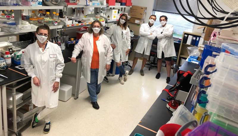 A group of students and faculty wearing masks and lab coats stand in a lab for a photo.
