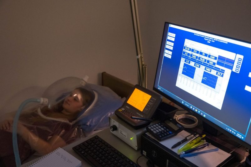 A person laying down in a bed with a scientific measuring instrument above them while a computer illuminates in darkness showing the results.